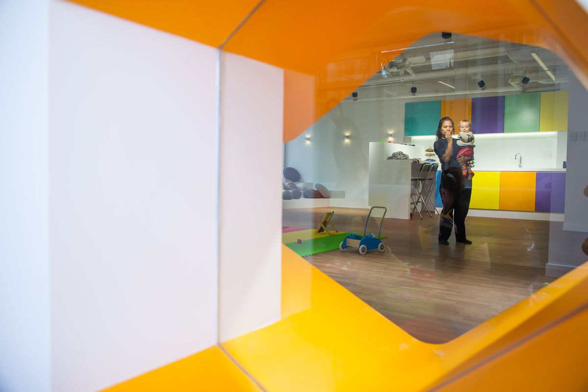 View through an orange framed opening into a bright early-learning room with colourful cabinets, a play mat and a carer holding a child