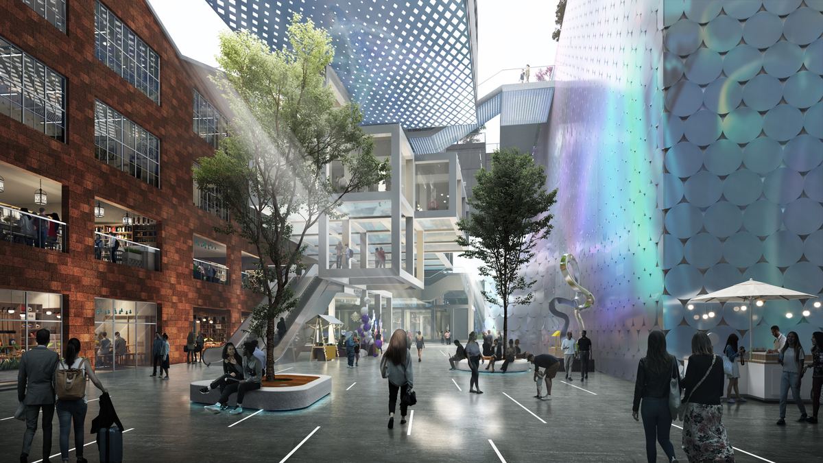 An open-air retail street between a converted brick warehouse and an iridescent facade, with trees, seating, market stalls and people strolling