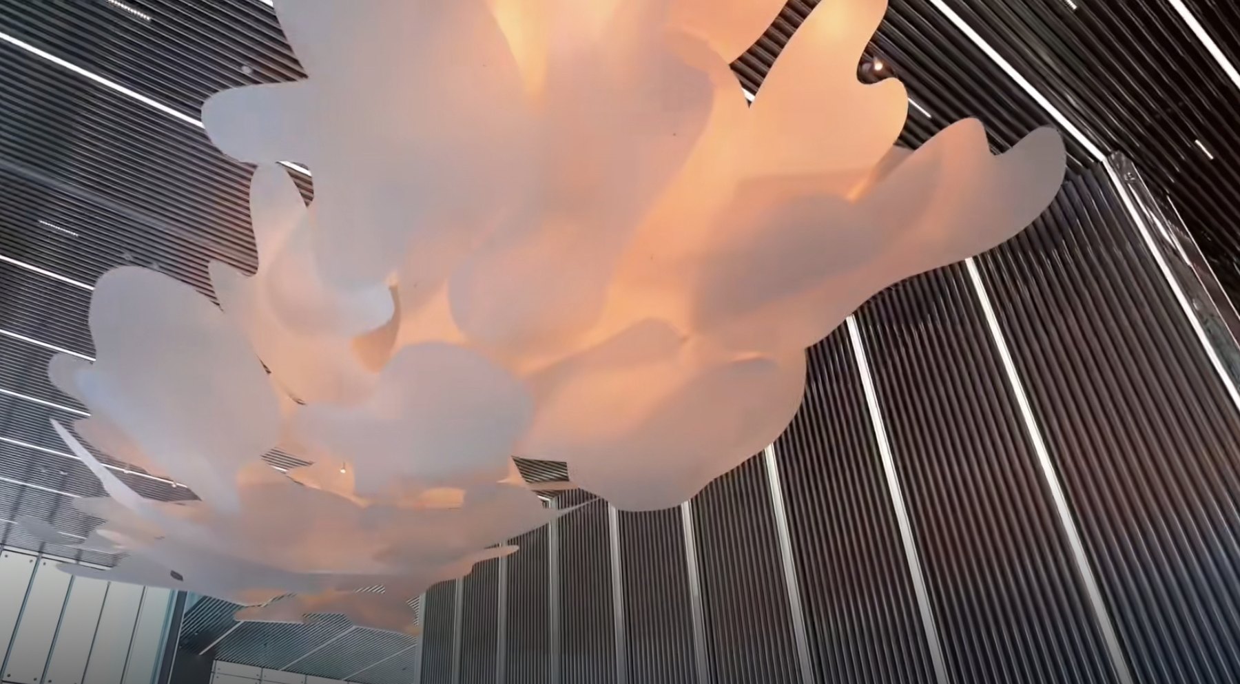 Looking up into the translucent cloud sculpture, its layered petals glowing warm against the linear steel ceiling and walls