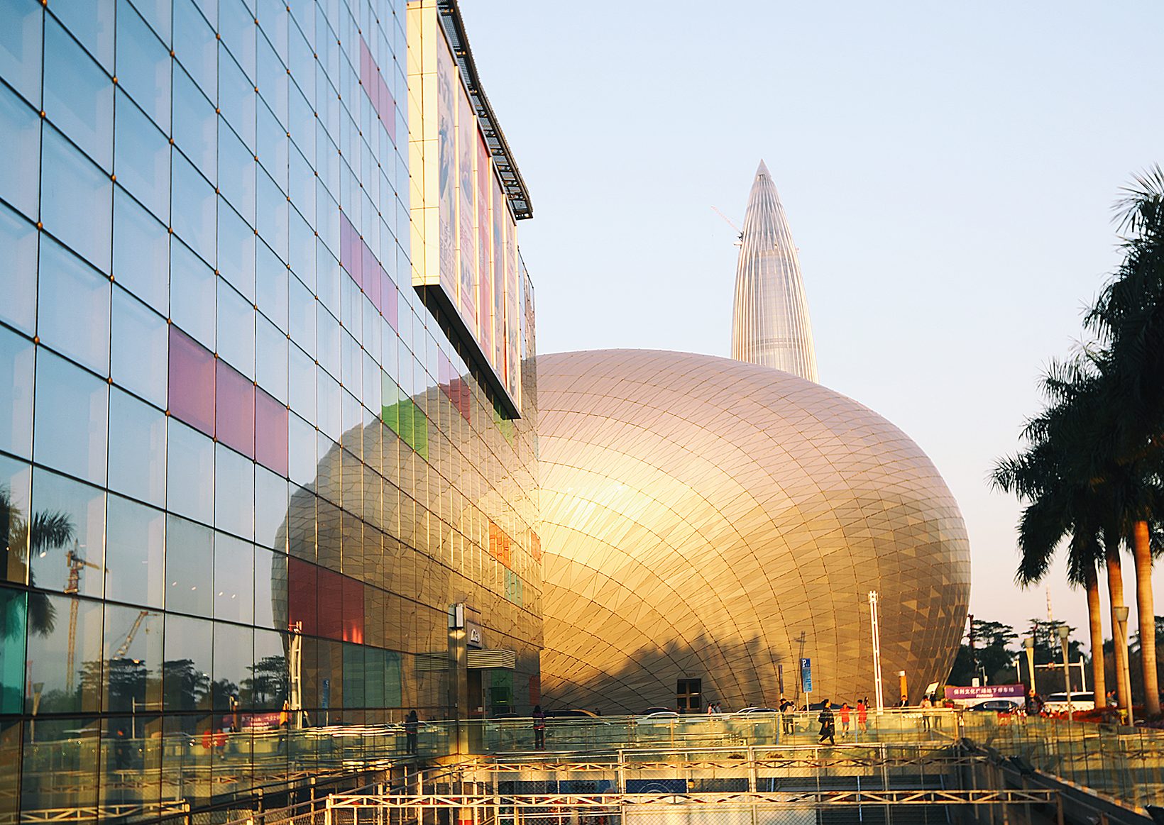 The golden oval of the Poly Theatre beside the glass wall of the commercial centre, palms and a tower beyond