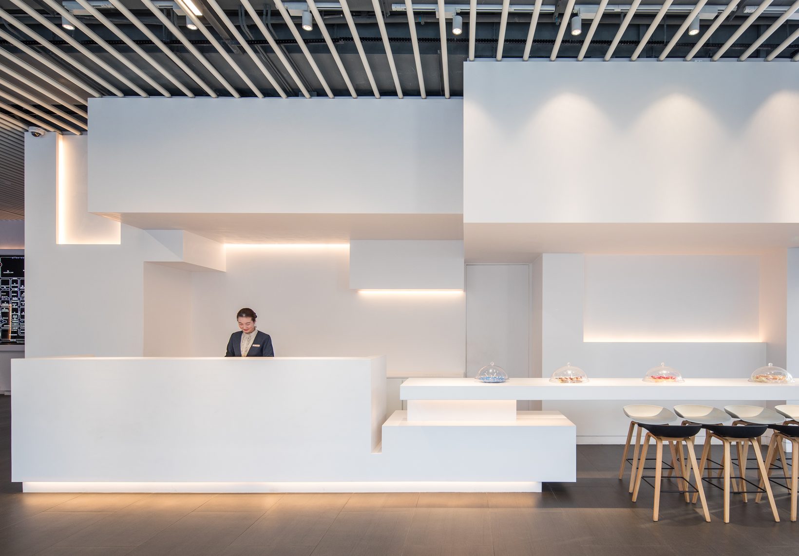 A white feature wall of stacked rectilinear volumes, lit from recessed slots, wrapping a reception desk and a bar with stools