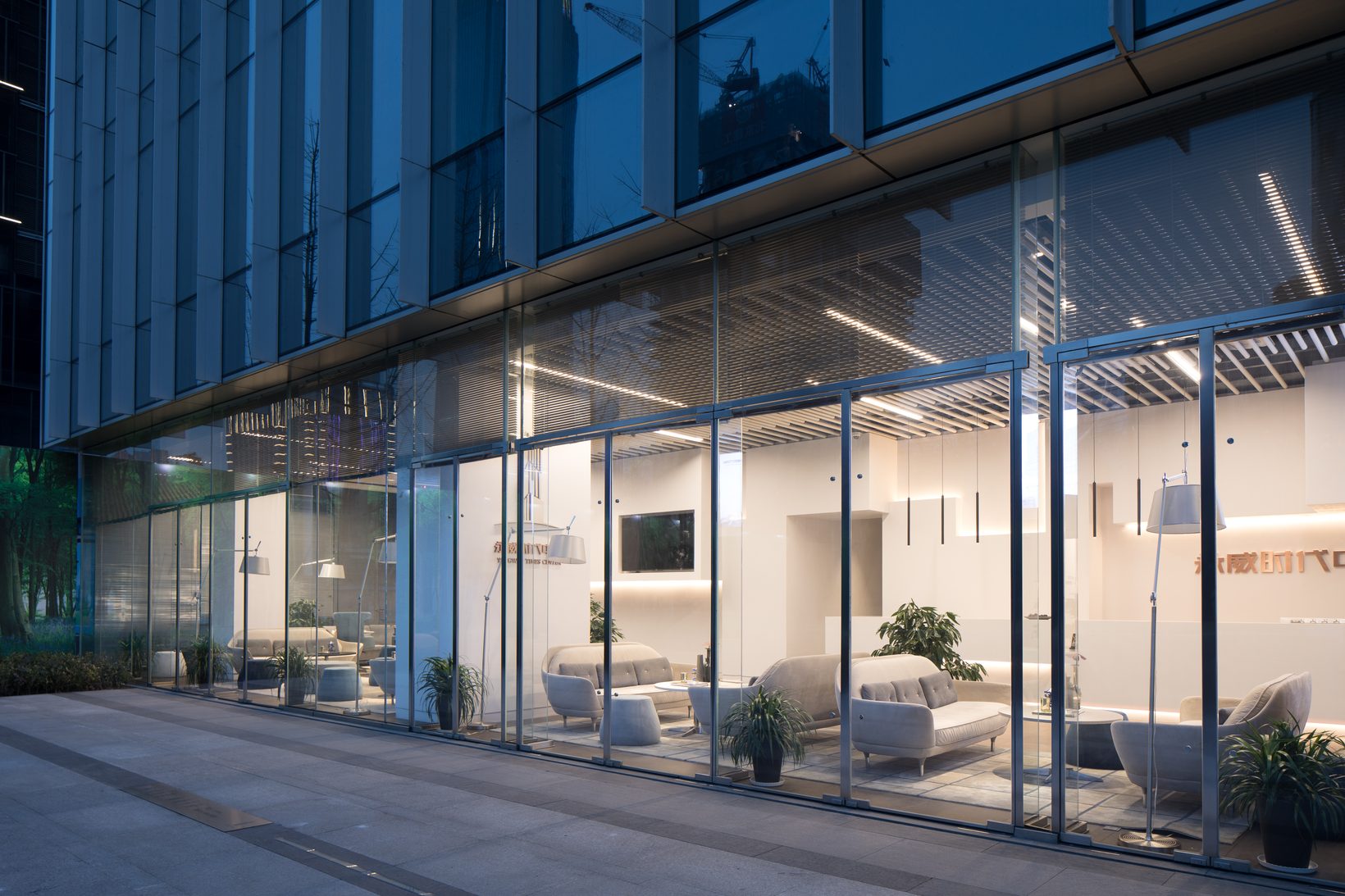 The sales centre at dusk, its glass front lit from within, lounge and white wall visible from the street